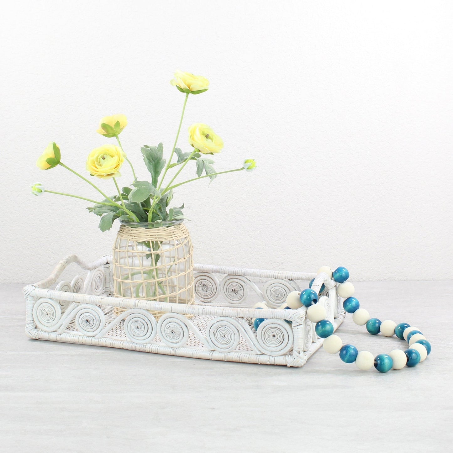 Whitewash rattan tray styled with yellow ranunculus flowers and blue decorative beads on a neutral tabletop.