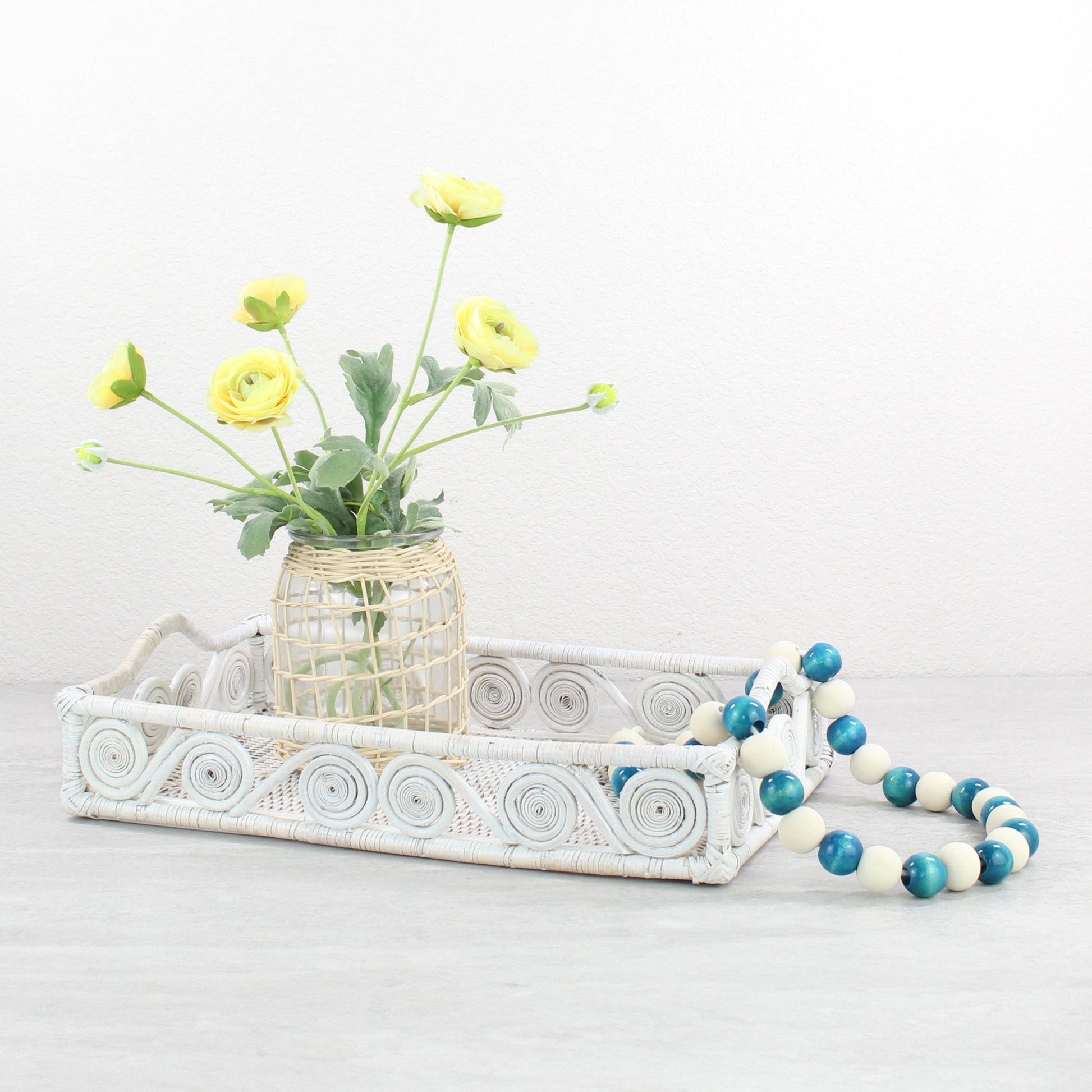 Whitewash rattan tray styled with yellow ranunculus flowers and blue decorative beads on a neutral tabletop.