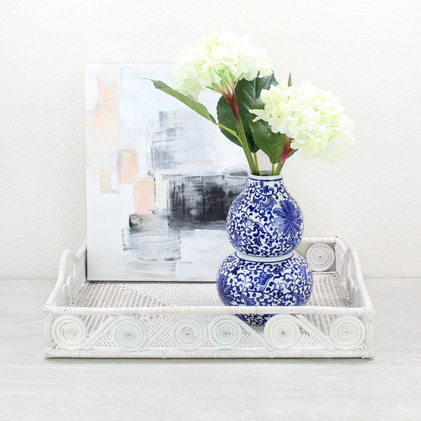 Whitewash rattan tray with swirl edge holding a blue floral vase of hydrangeas in front of a modern abstract painting.