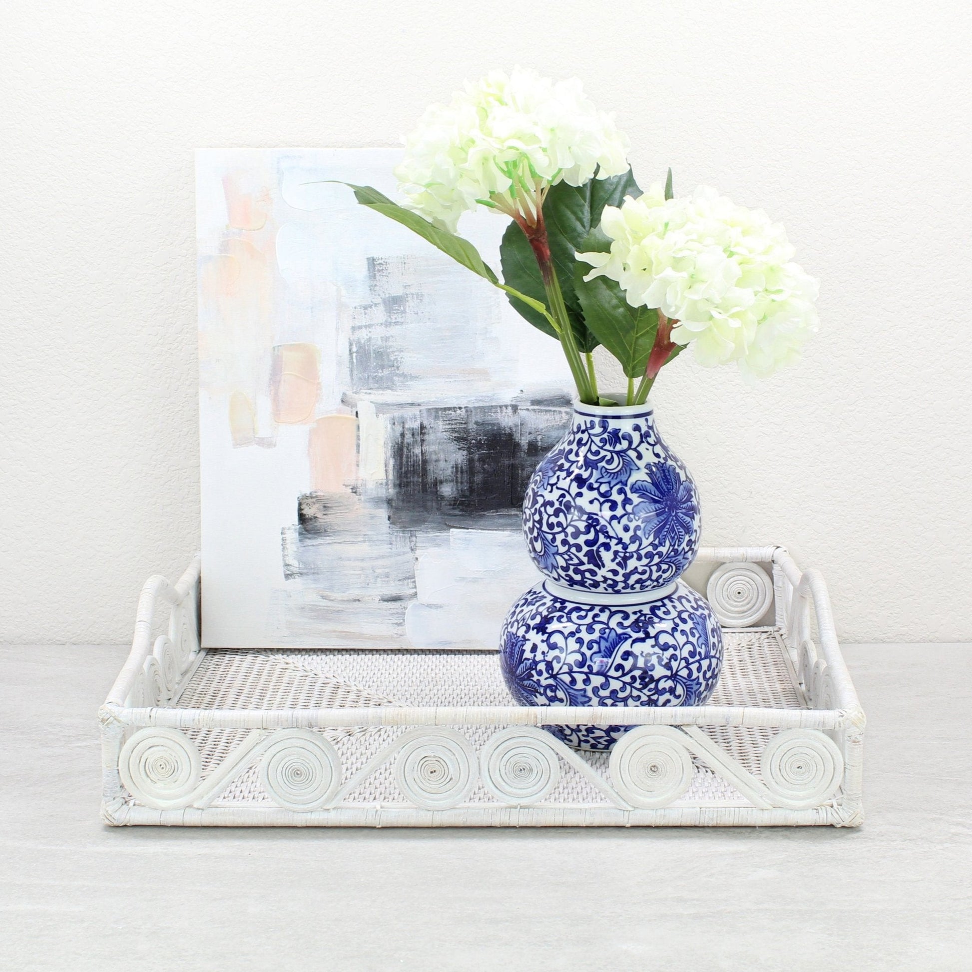 Whitewash rattan tray with swirl edge holding a blue floral vase of hydrangeas in front of a modern abstract painting.