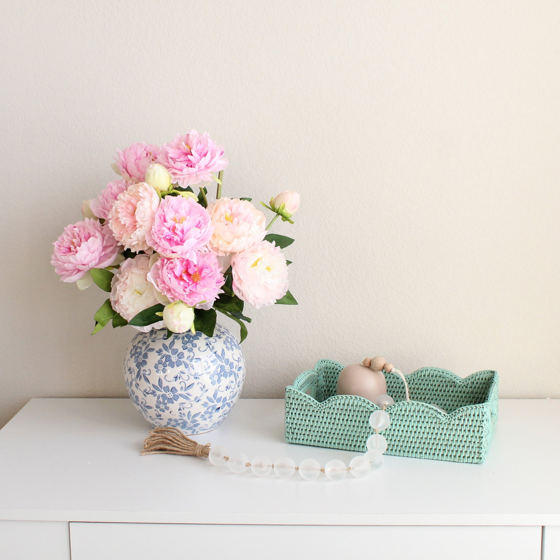 Sage Leaf Green rattan tray with scalloped edge styled on a white table beside a blue floral vase of pink peonies and a bead garland.