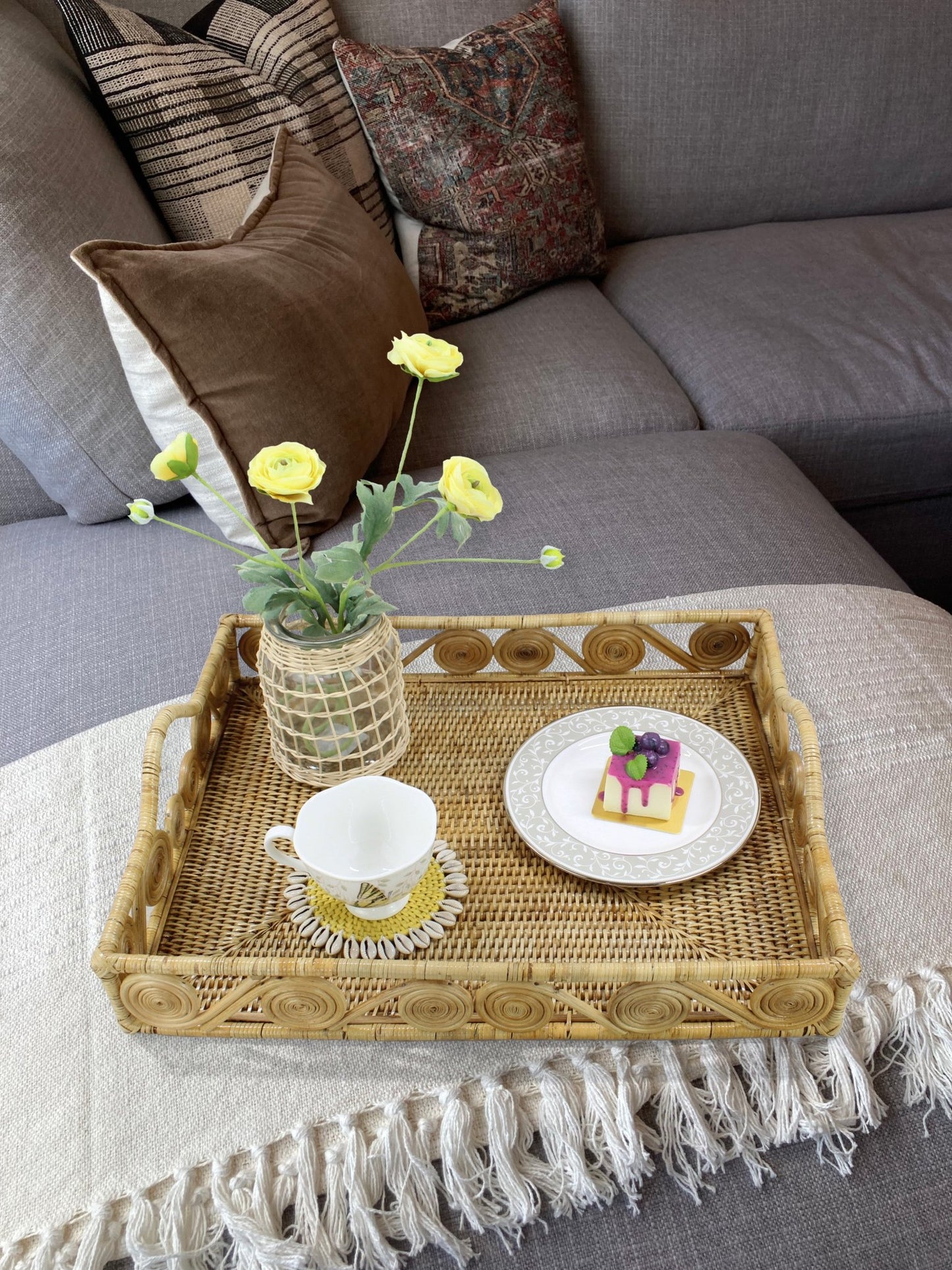 Natural rectangular rattan tray with swirl design styled on a gray sofa coffee table, holding yellow flowers, a teacup, and dessert plate.