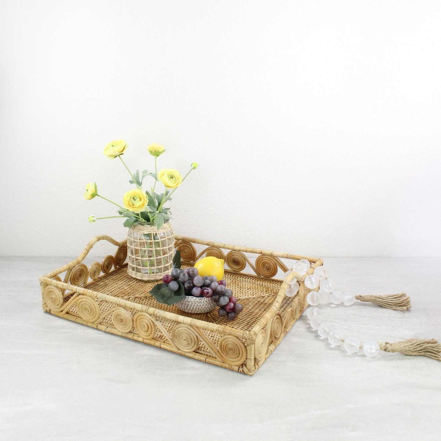 Natural rectangular rattan tray with swirl edge, decorated with yellow flowers, a lemon, grapes, and a frosted bead garland for rustic décor styling.