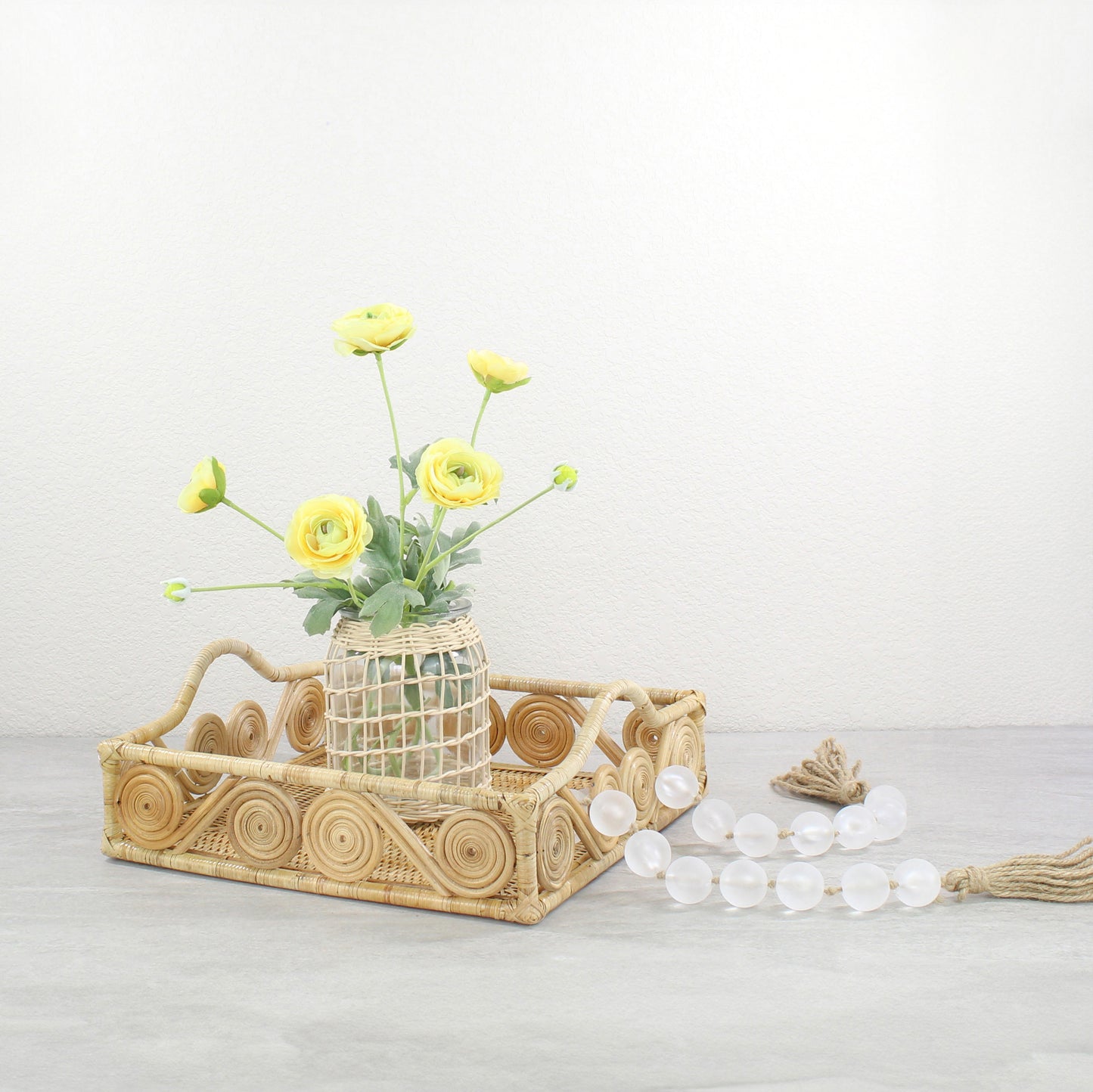 Natural rattan tray styled with yellow ranunculus flowers in a woven vase and frosted bead garland.
