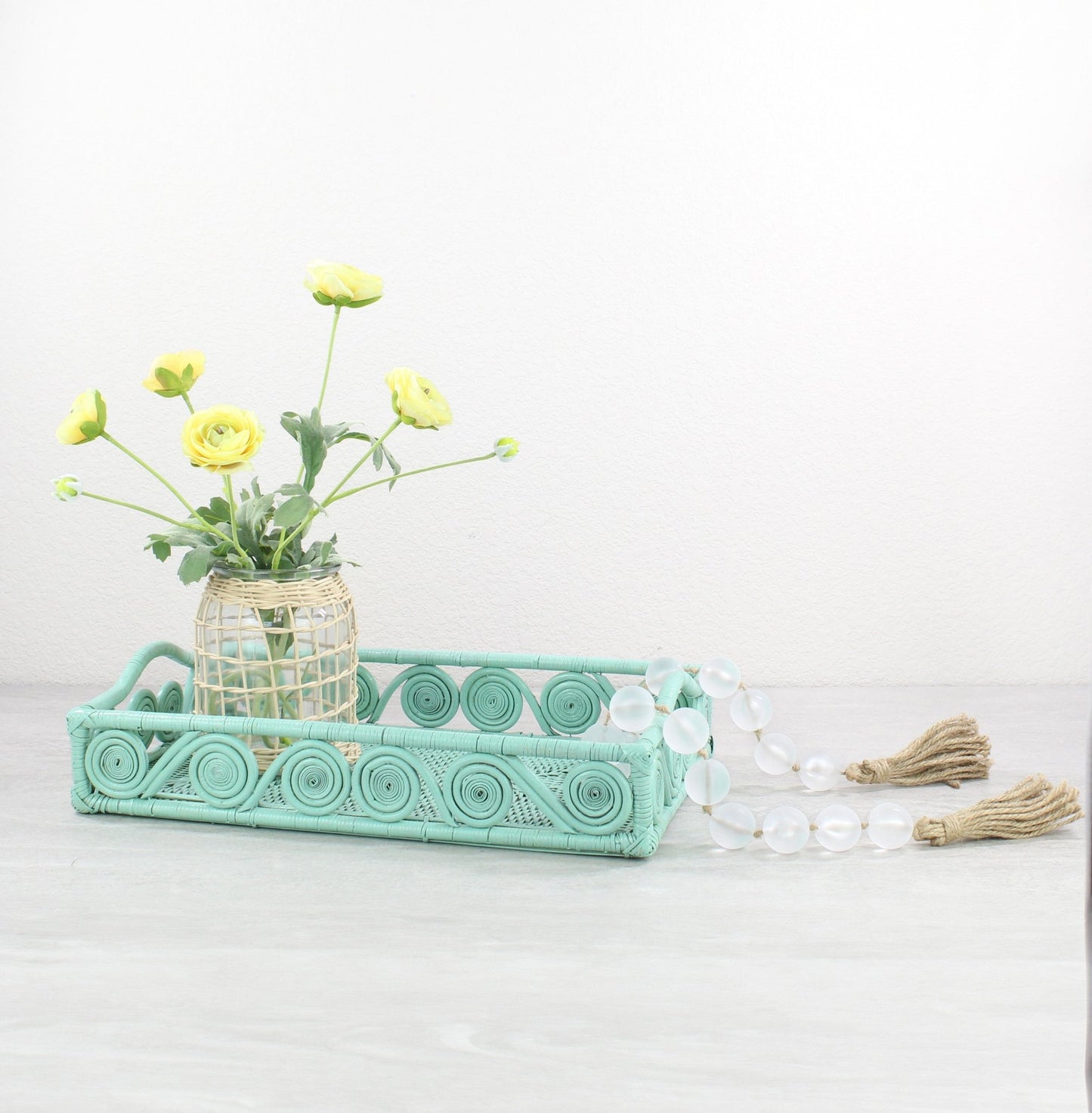 Sage leaf green swirly rattan tray styled with yellow ranunculus flowers and glass decorative beads on a neutral tabletop.