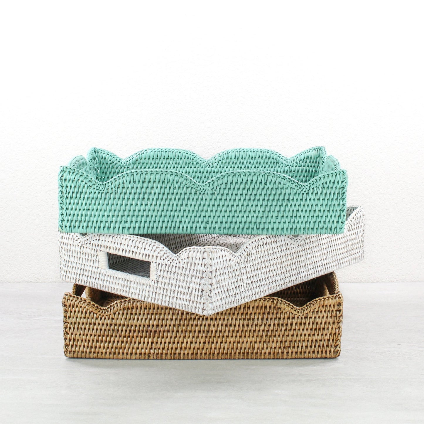 Handwoven scalloped edge rattan trays with handles in turquoise, whitewash, and honey brown stacked together.
