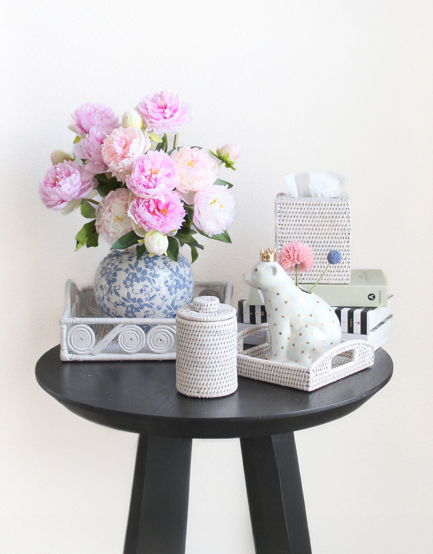 Whitewash rattan trays, canister, and tissue box cover styled with pink peonies, books, and décor on a black accent table