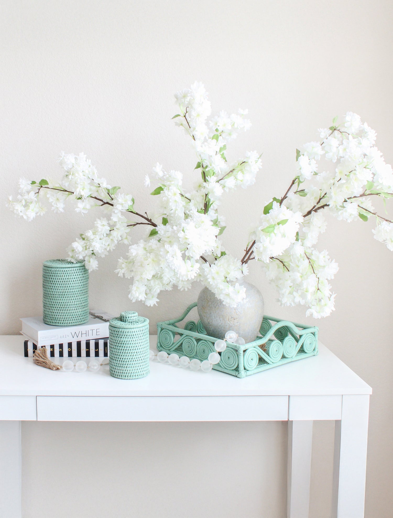 Sage green rattan tray and canisters styled with white floral arrangement on white table, showcasing elegant handmade home décor.