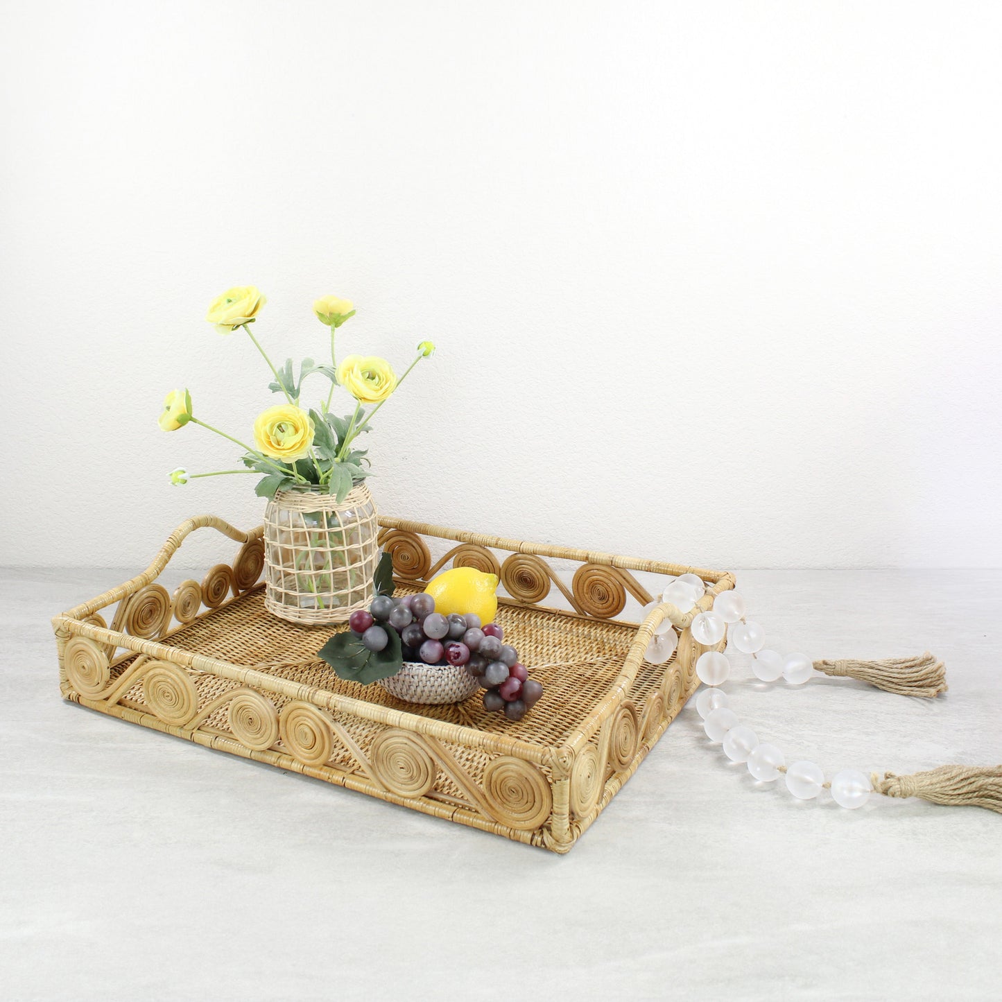 Natural rattan tray decorated with yellow ranunculus flowers, a lemon, grapes, and frosted bead garland.