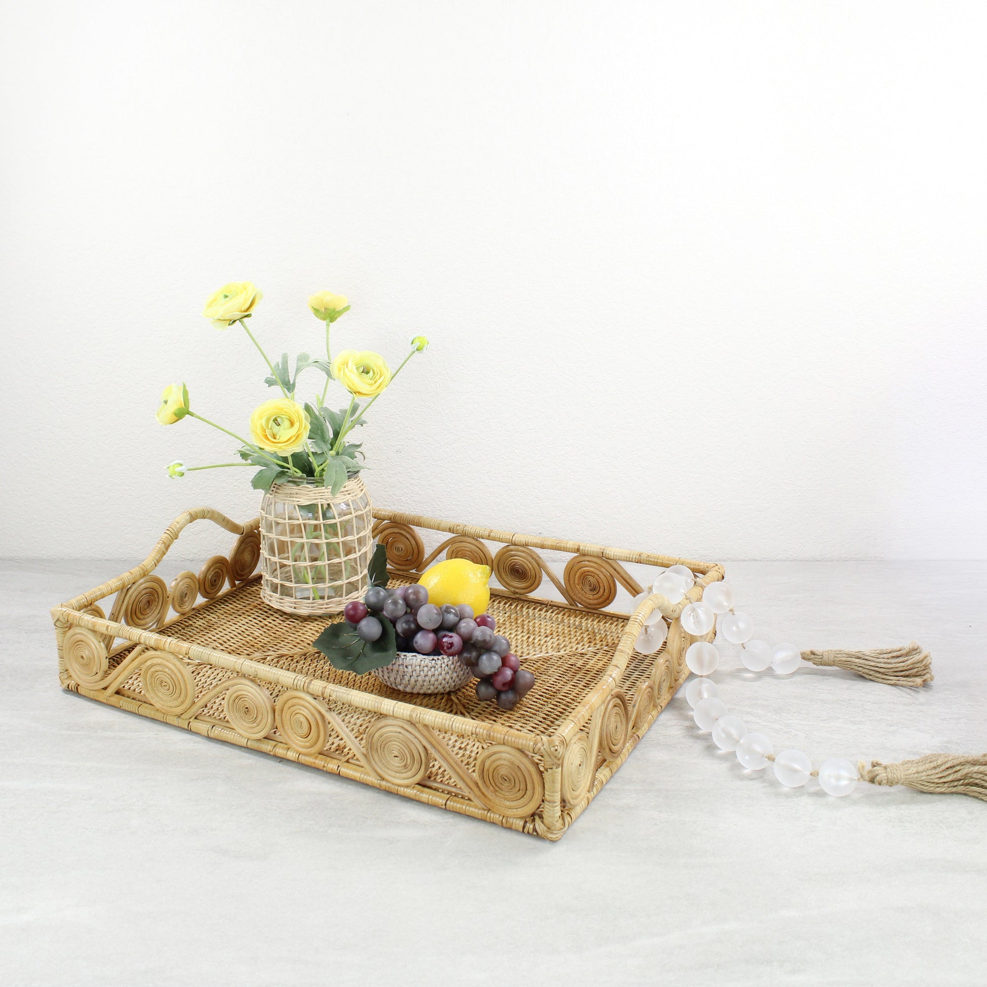 Natural rattan tray decorated with yellow ranunculus flowers, a lemon, grapes, and frosted bead garland.