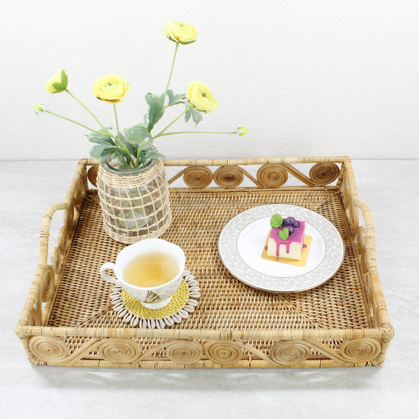 Natural rattan tray styled as a tea serving set with yellow flowers, teacup on seashell coaster, and dessert plate.