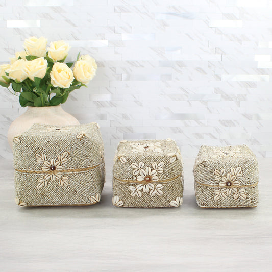 Three handcrafted beige beaded storage boxes with cowrie shell floral designs displayed beside a cream vase with roses on a neutral background.