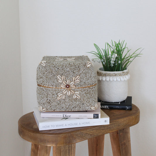 Handcrafted beige beaded and cowrie shell decorative box displayed on a wooden stool, styled with books and a white woven planter with greenery in a modern coastal home setting.