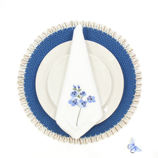 Blue rattan placemat with seashell trim, white plate, and floral napkin styled on a white dining table for coastal decor.