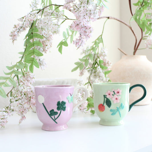 Pink and green handpainted stoneware garden mugs with floral patterns, displayed among pink flowers and leafy greenery.