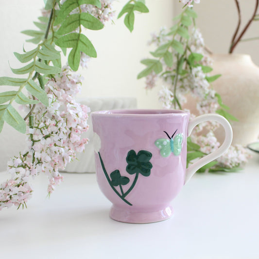 Pink handpainted stoneware garden mugs with floral patterns, displayed on table among pink flowers and leafy greenery.