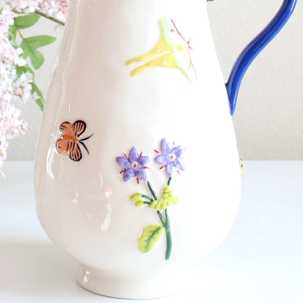 White stoneware pitcher with hand-painted garden-inspired motifs including flowers and butterflies, artisan glazed ceramic jug, close view
