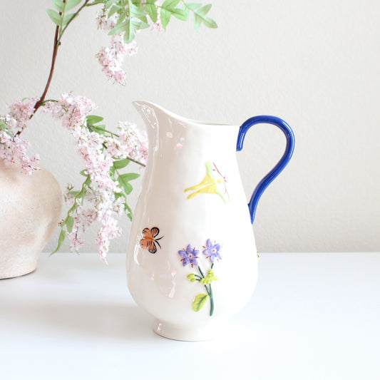 White stoneware pitcher with hand-painted garden-inspired motifs including flowers, butterflies, and ladybugs, artisan glazed ceramic jug