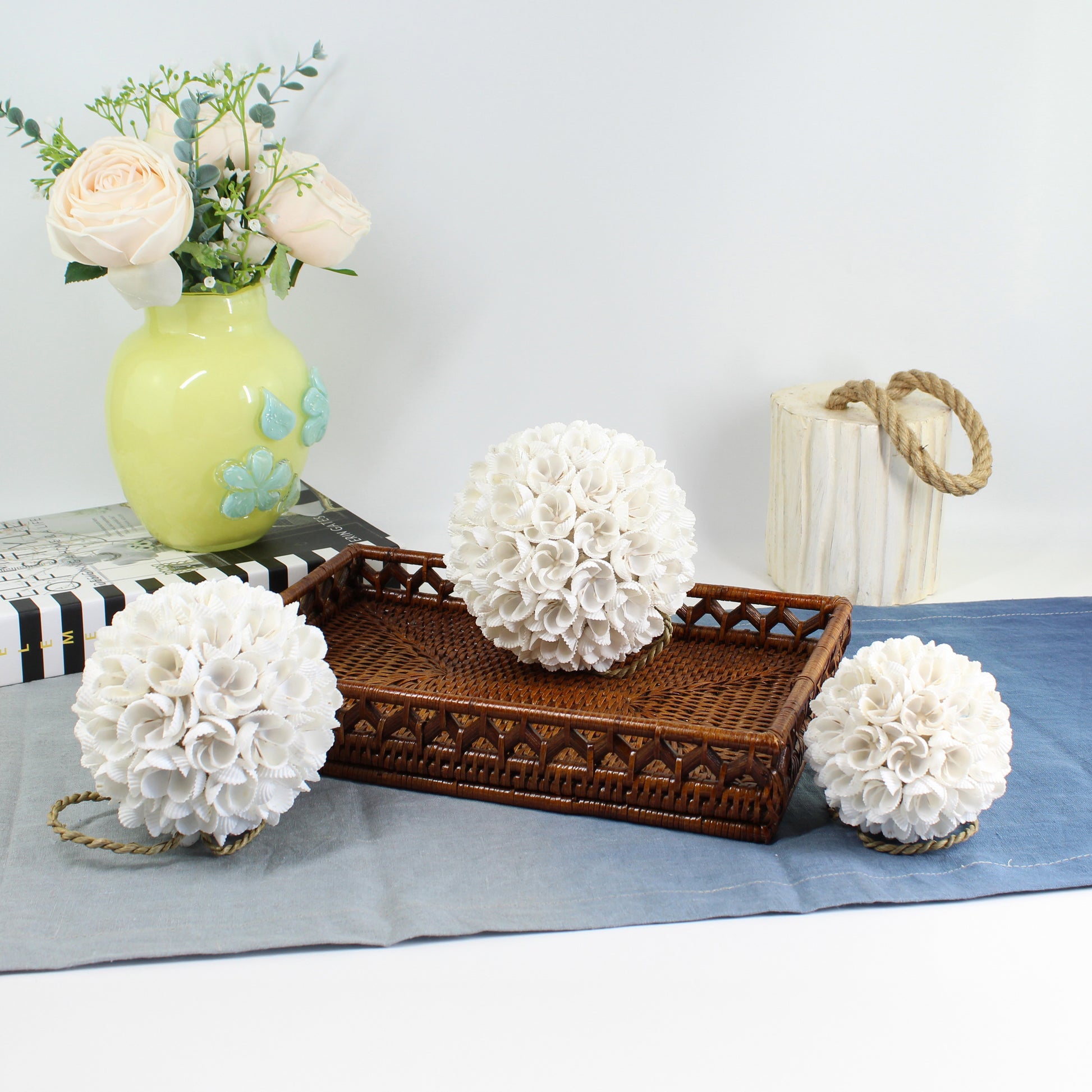 Eco Handcraft handmade seashell blossom ball décor in three sizes, styled with a rattan tray, yellow vase with flowers, and soft blue table runner.