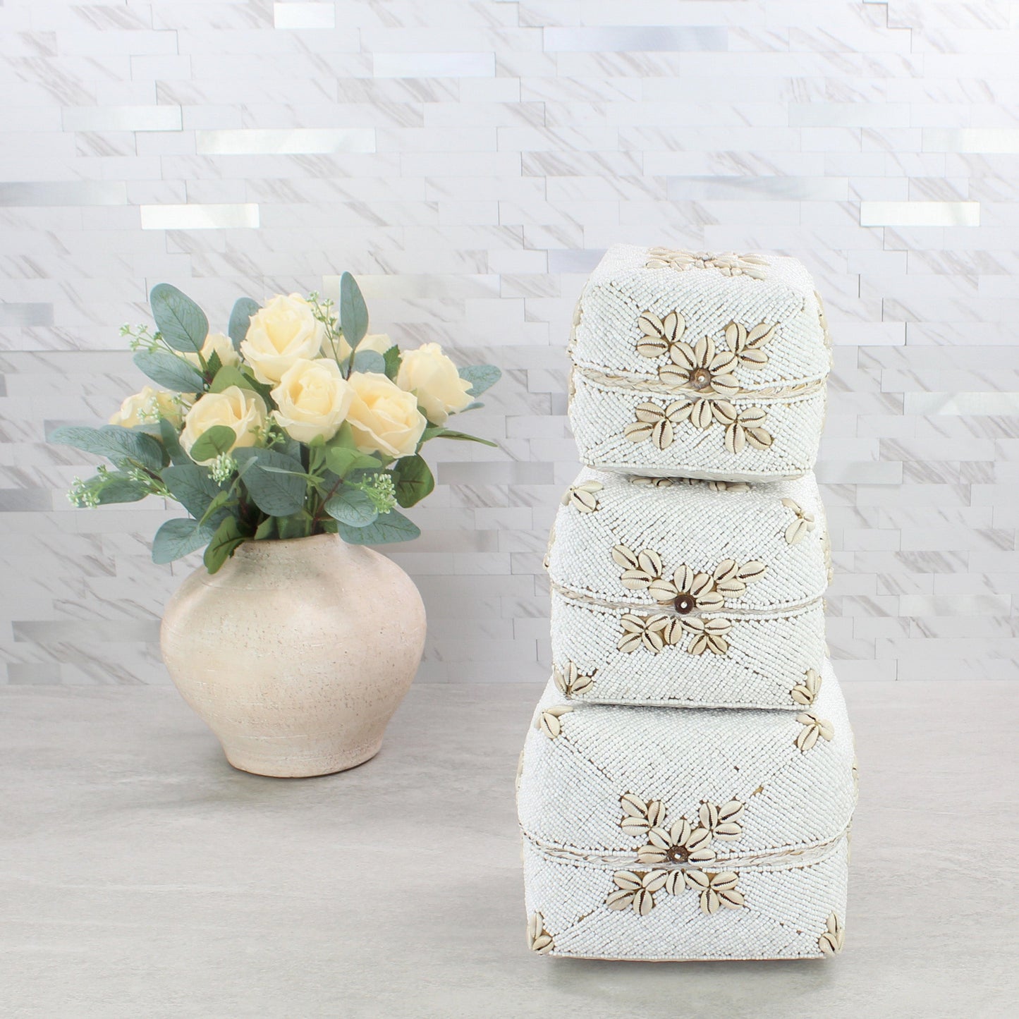 Handmade white beaded cowrie shell storage boxes stacked beside a beige vase with roses on marble background.