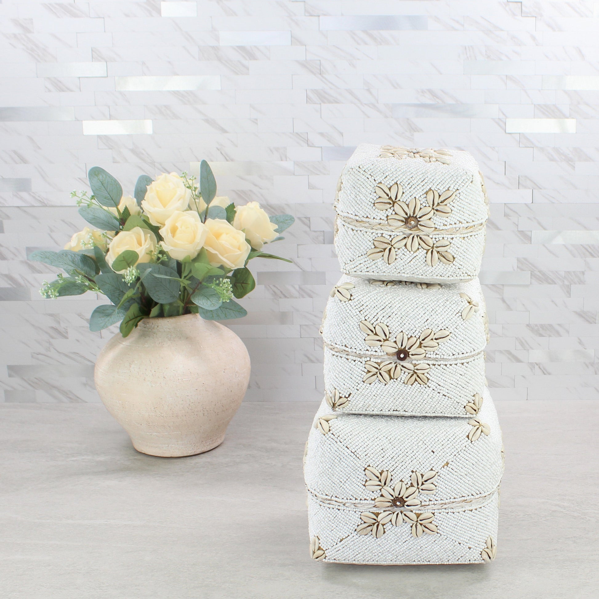Handmade white beaded cowrie shell storage boxes stacked beside a beige vase with roses on marble background.