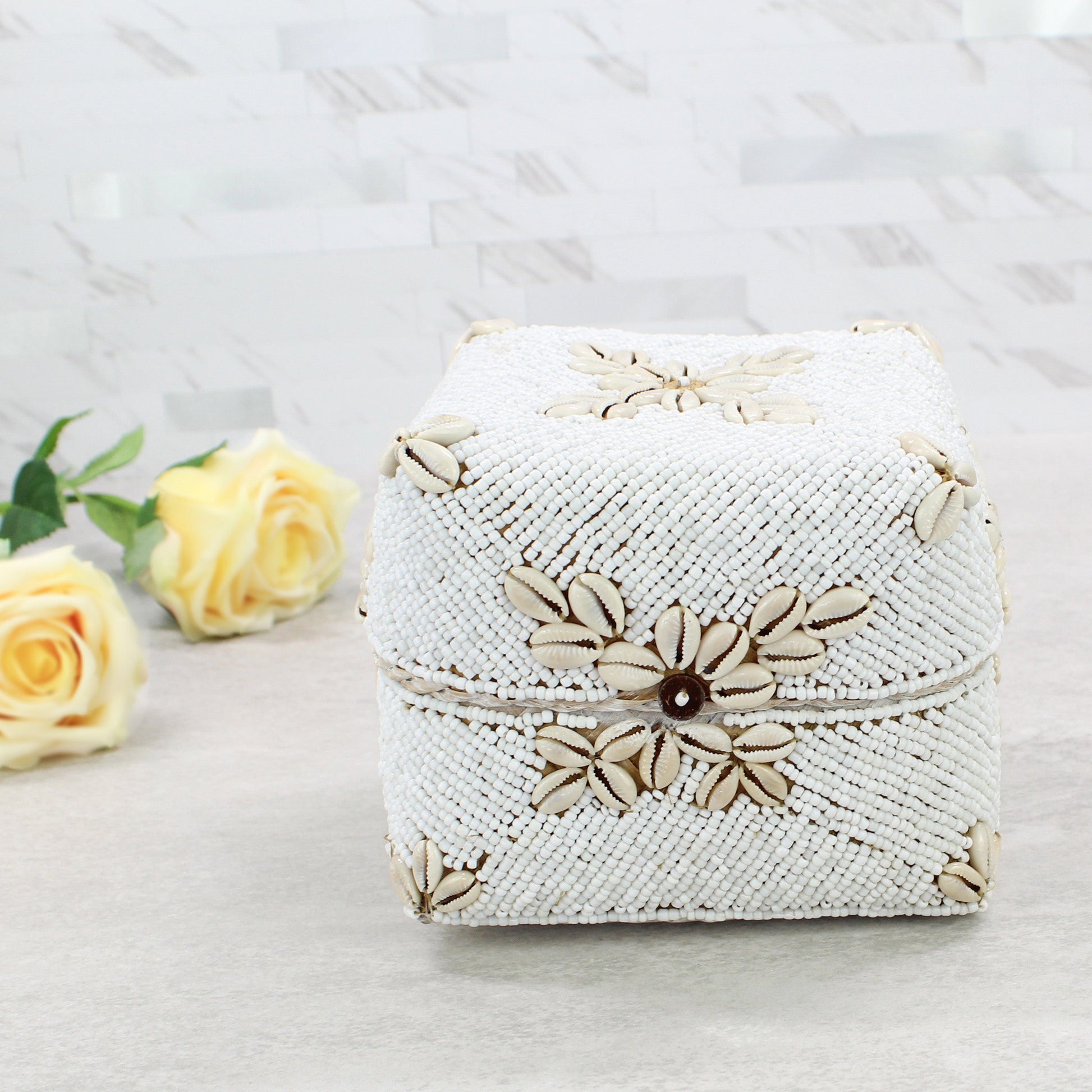 Medium handmade white beaded cowrie shell decorative storage box displayed with yellow roses on neutral countertop.