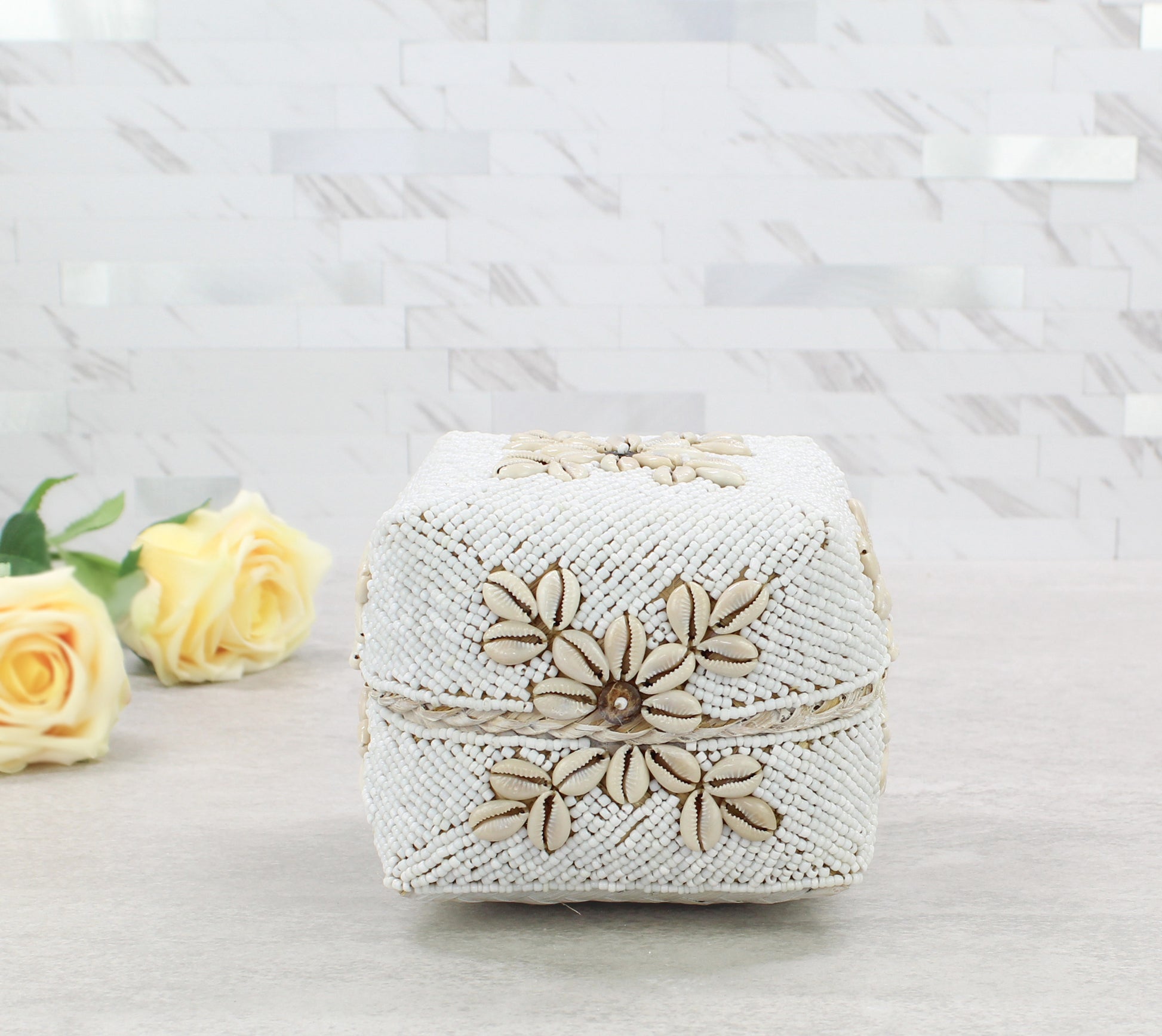 Small handmade white beaded cowrie shell decorative storage box displayed with yellow roses on neutral countertop.