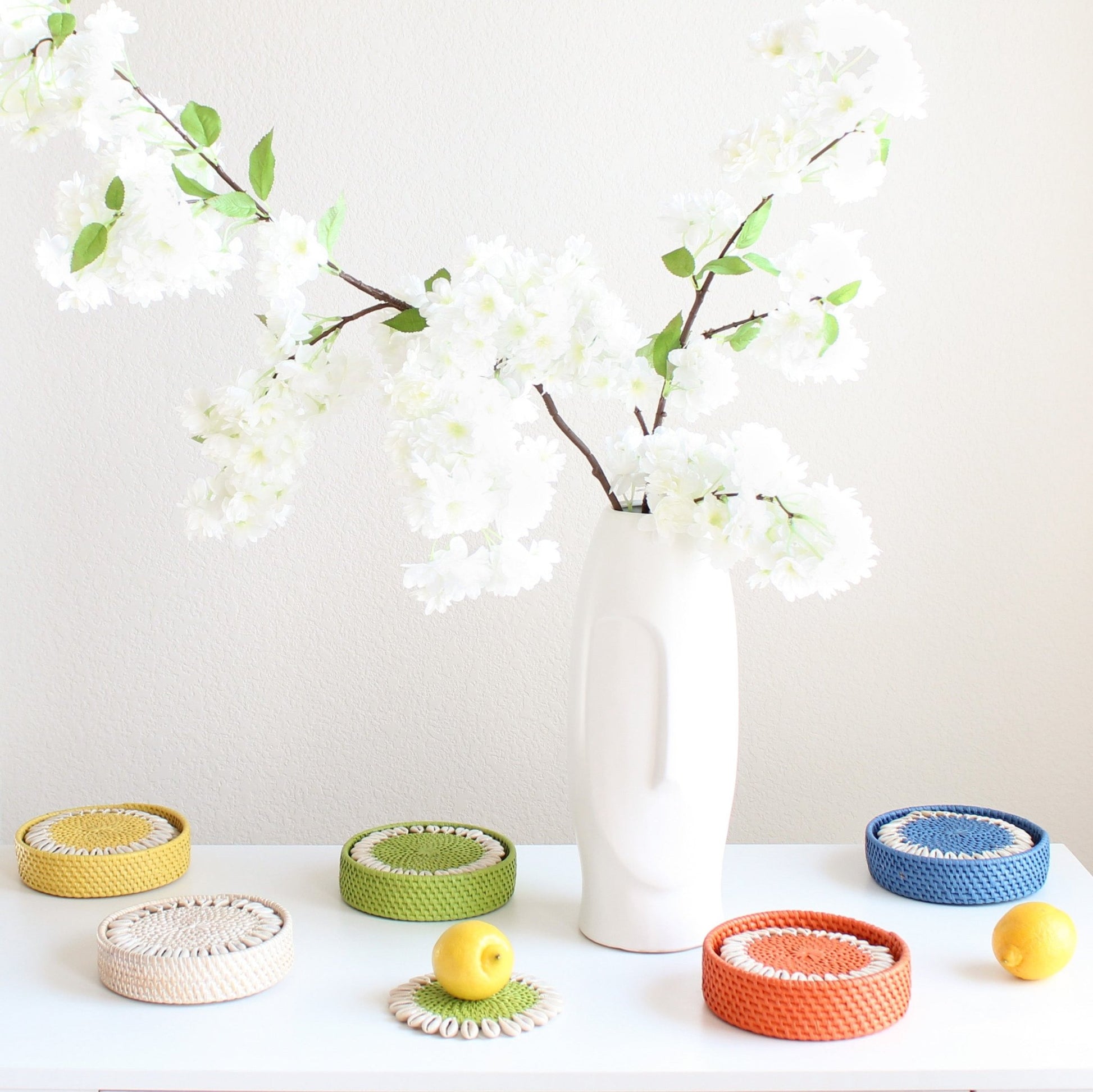 Colorful rattan coasters with seashell trim styled near vase with flowers and citrus fruits for vibrant coastal table décor.