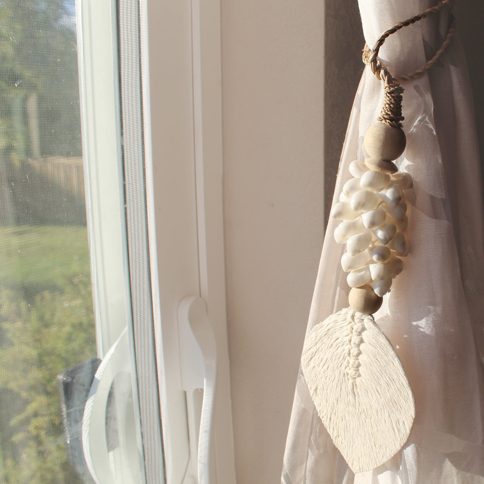 White seashell tassel curtain tieback with macramé leaf design hanging by sunlit window in neutral room