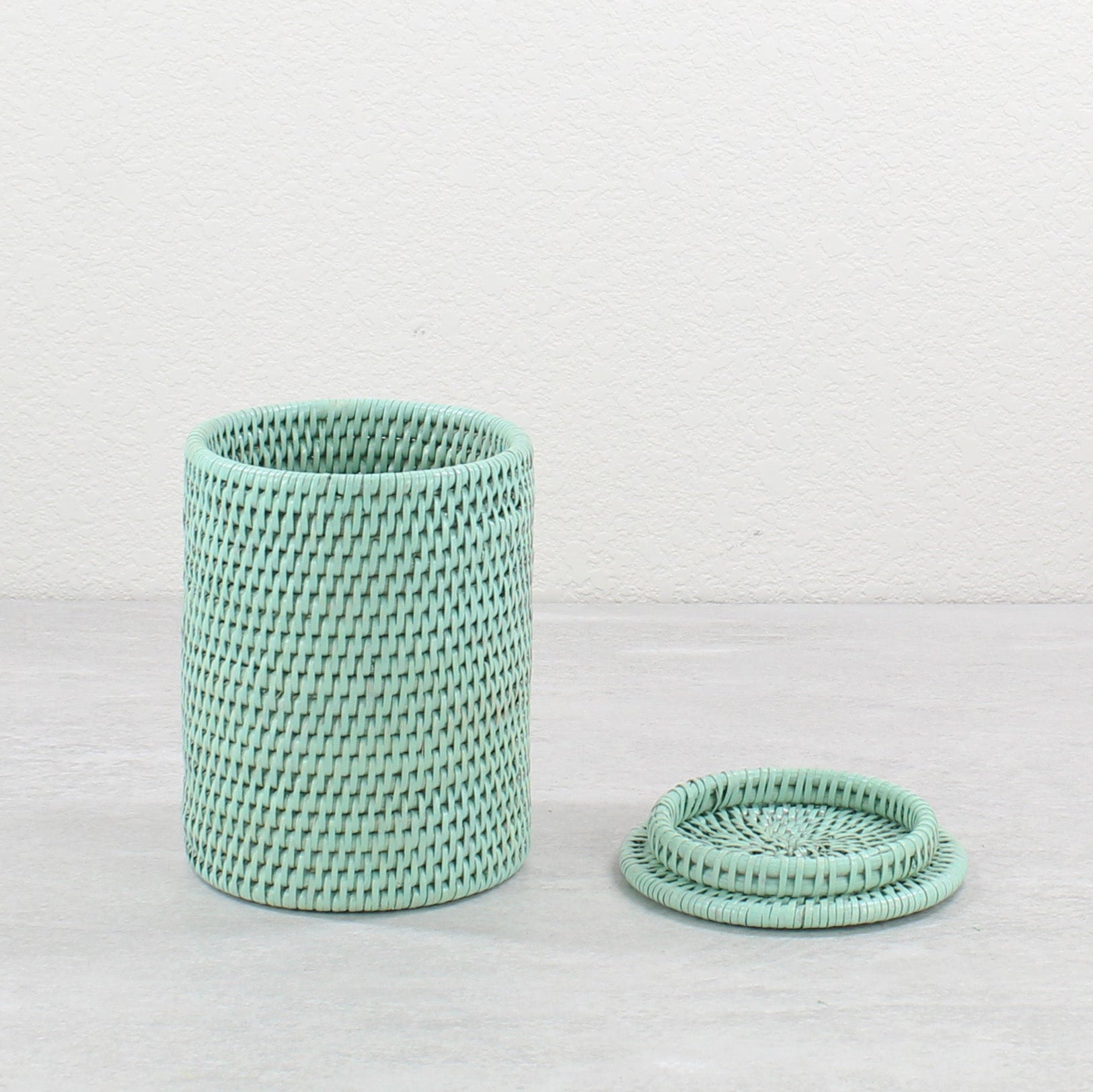 Sage green handwoven rattan canister, lid placed beside on light gray surface.