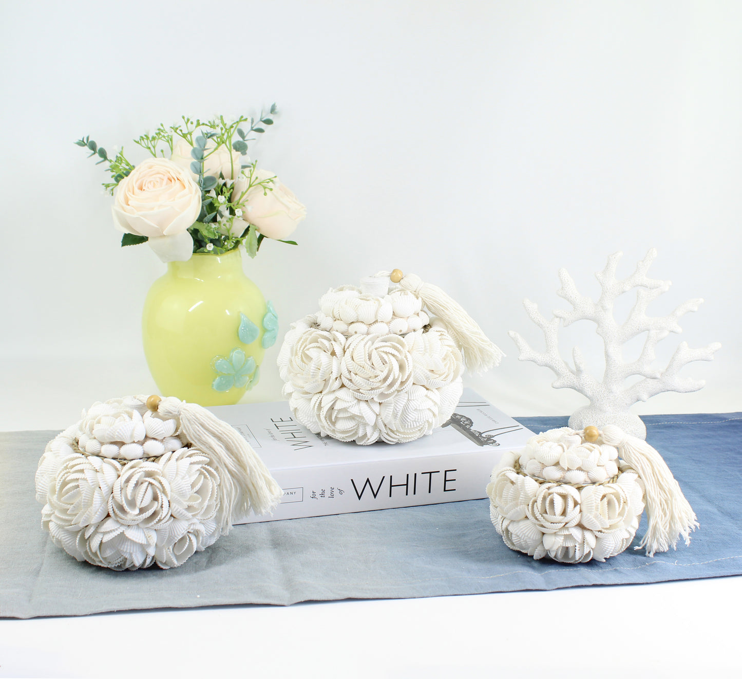 Set of three handmade rose seashell storage boxes with white tassels, displayed with roses in a green vase, artisan coastal home décor with blue table runner.