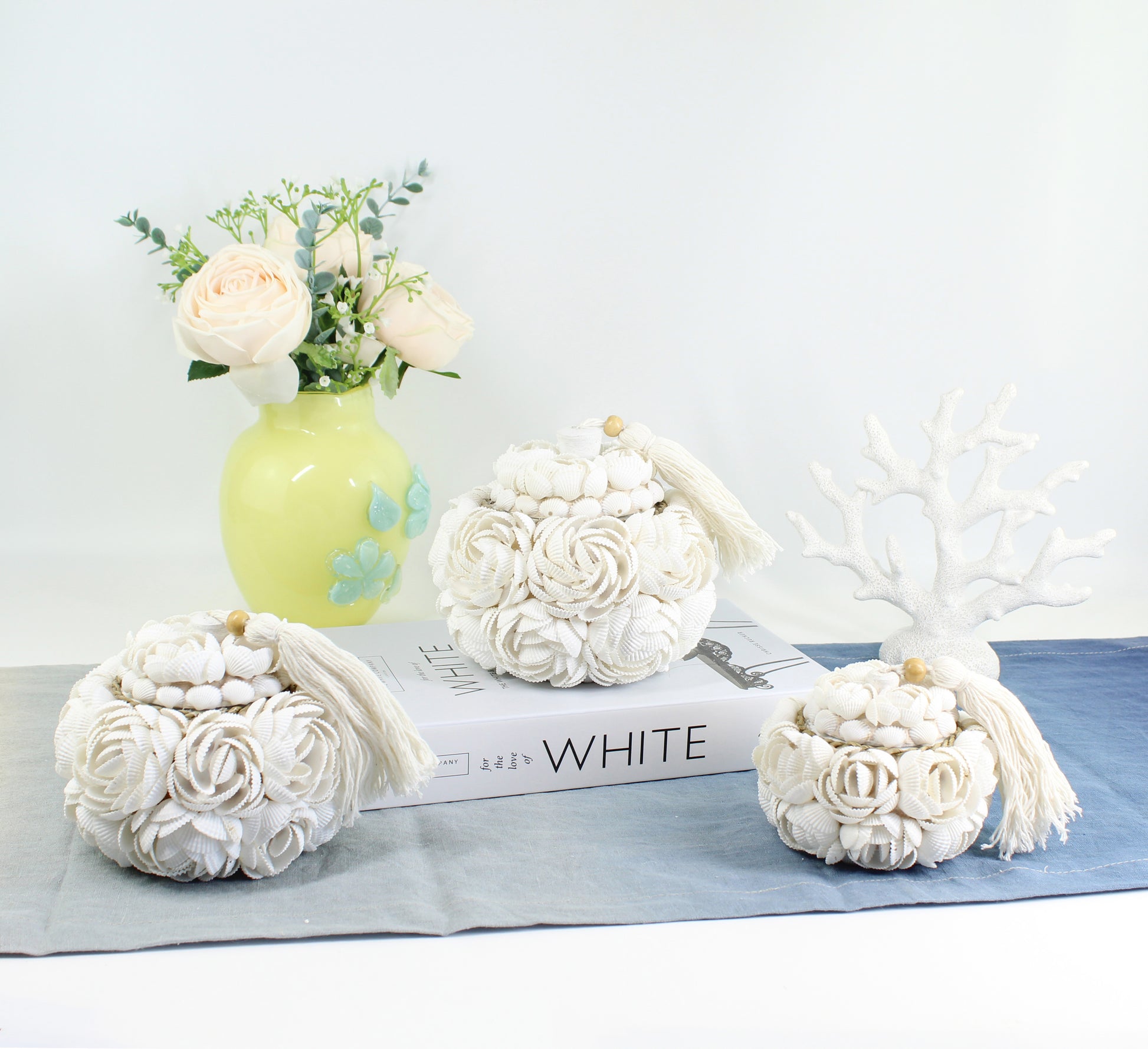 Set of three handmade rose seashell storage boxes with white tassels, displayed with roses in a green vase, artisan coastal home décor with blue table runner.