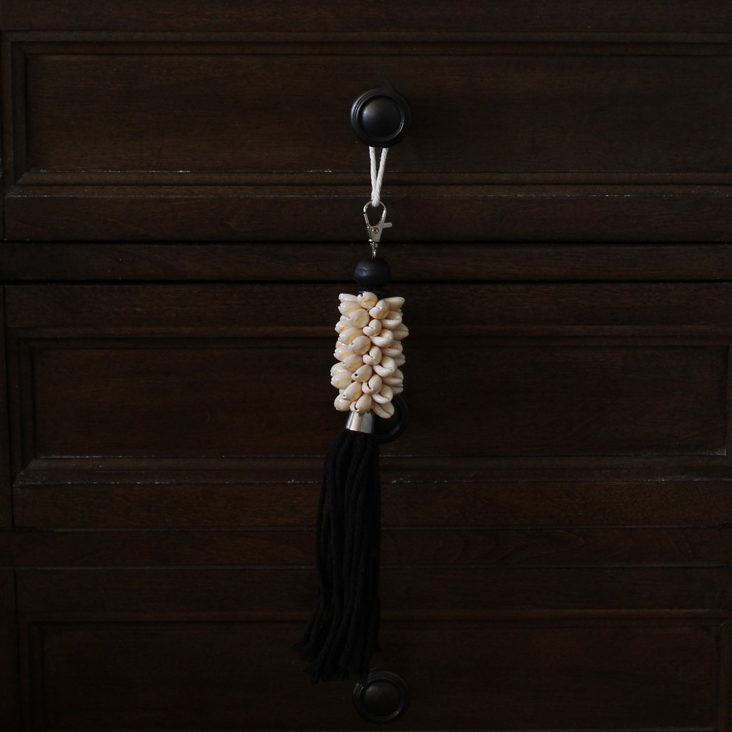 Handmade natural seashell key ring with black cotton tassel hanging from dark wood drawer knob, artisan boho accent.