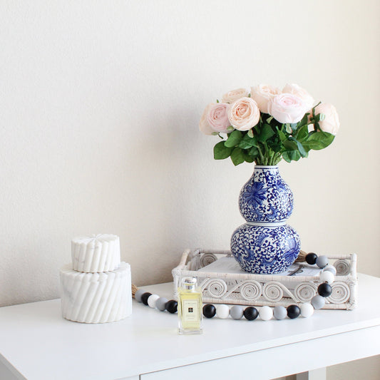White swirly rattan tray styled with blue floral vase, roses, and perfume on white table for chic modern home décor.