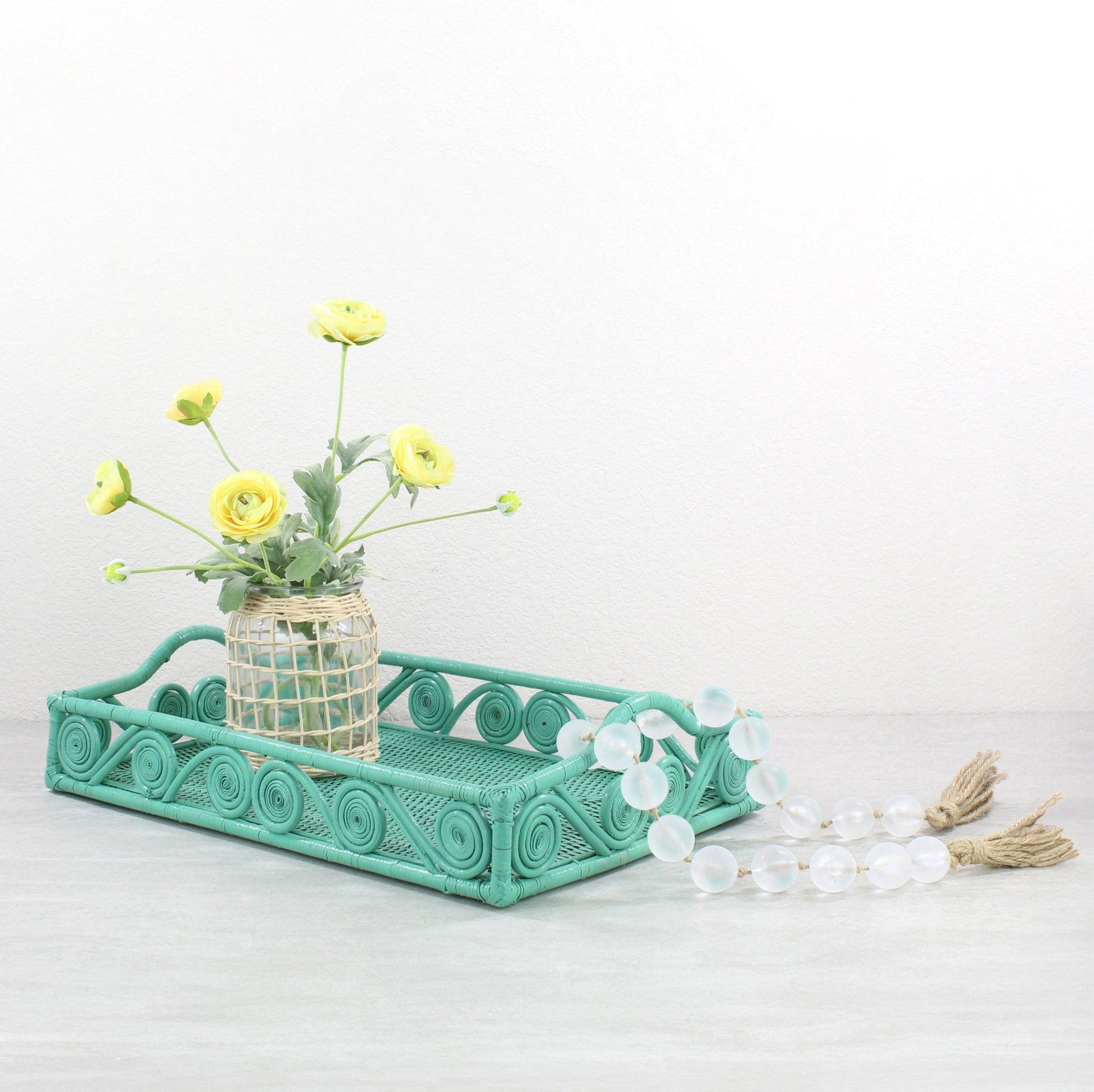 Turquoise swirly rattan tray styled with yellow flowers in a woven vase and glass bead garland on a gray tabletop.