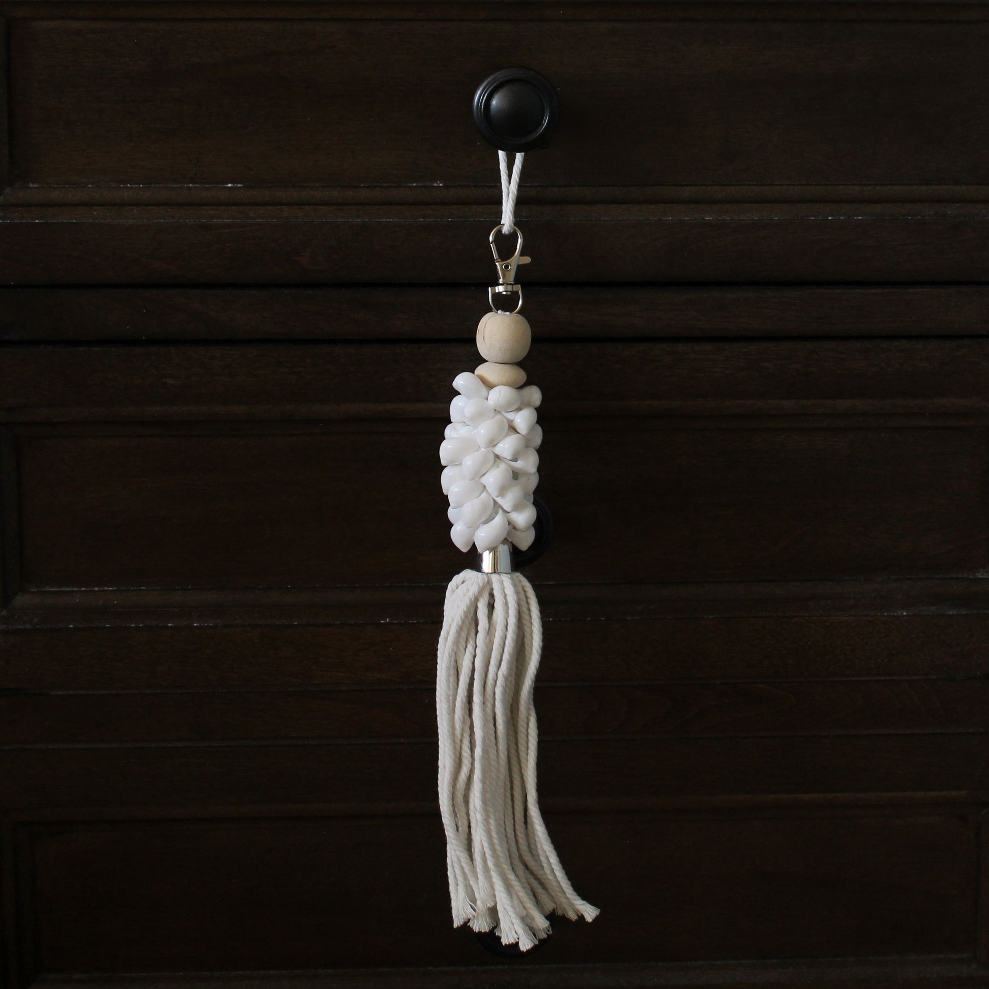 Gloss white seashell cluster key ring with white cotton tassel hanging from dark wood drawer, handmade coastal accessory.