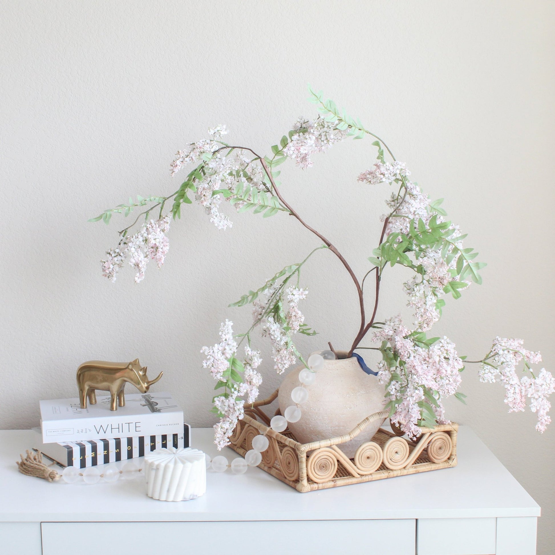 Natural swirly rattan tray styled on a white console table with a ceramic vase of blossoms, bead garland, and gold rhino figurine.