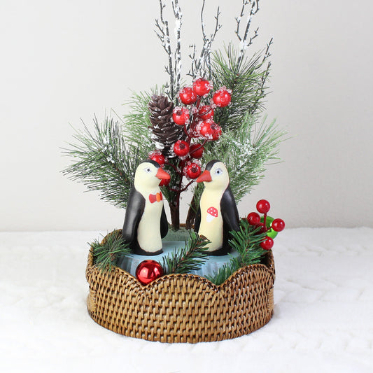 Holiday gift idea featuring hand-painted penguin couple figurines displayed on scalloped round honey brown rattan tray with festive décor accents