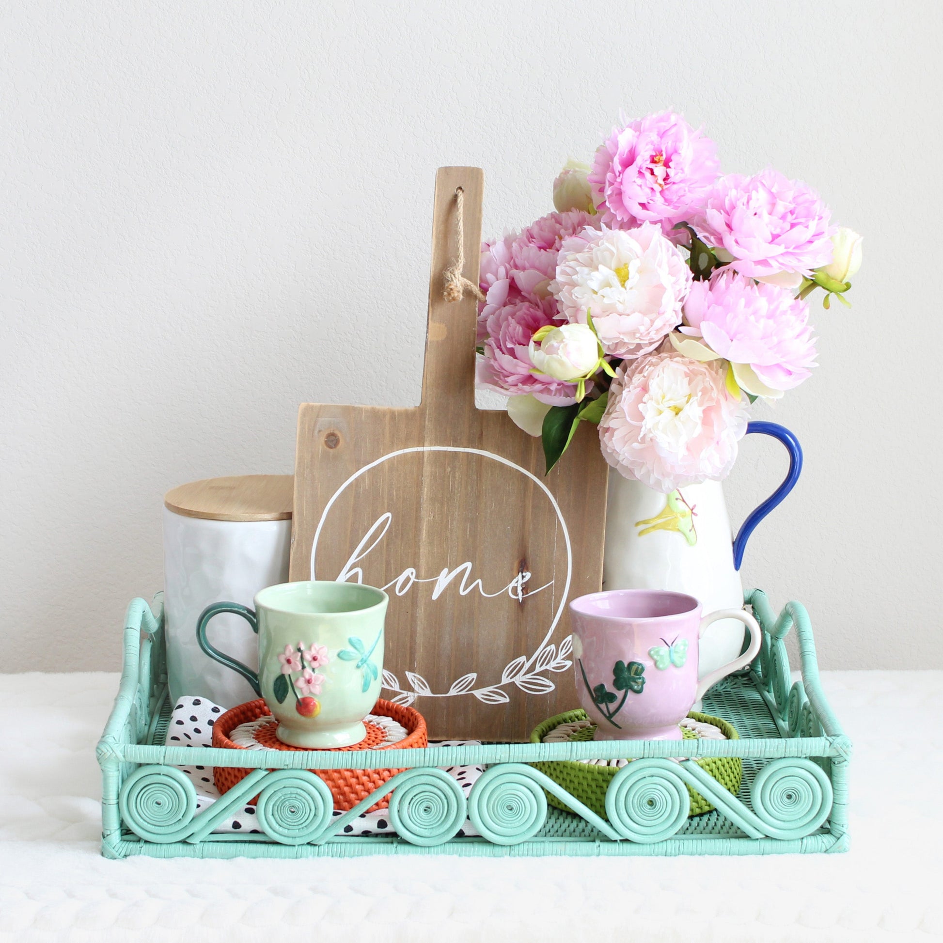 Housewarming gift set styled with handwoven sage leaf green rattan tray, seashell coasters, hand-painted stoneware mugs, and floral pitcher with pink flowers