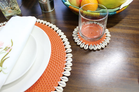Orange rattan coaster with seashell trim styled on a wooden dining table with glassware and matching seashell placemat for an elegant coastal-inspired tablescape