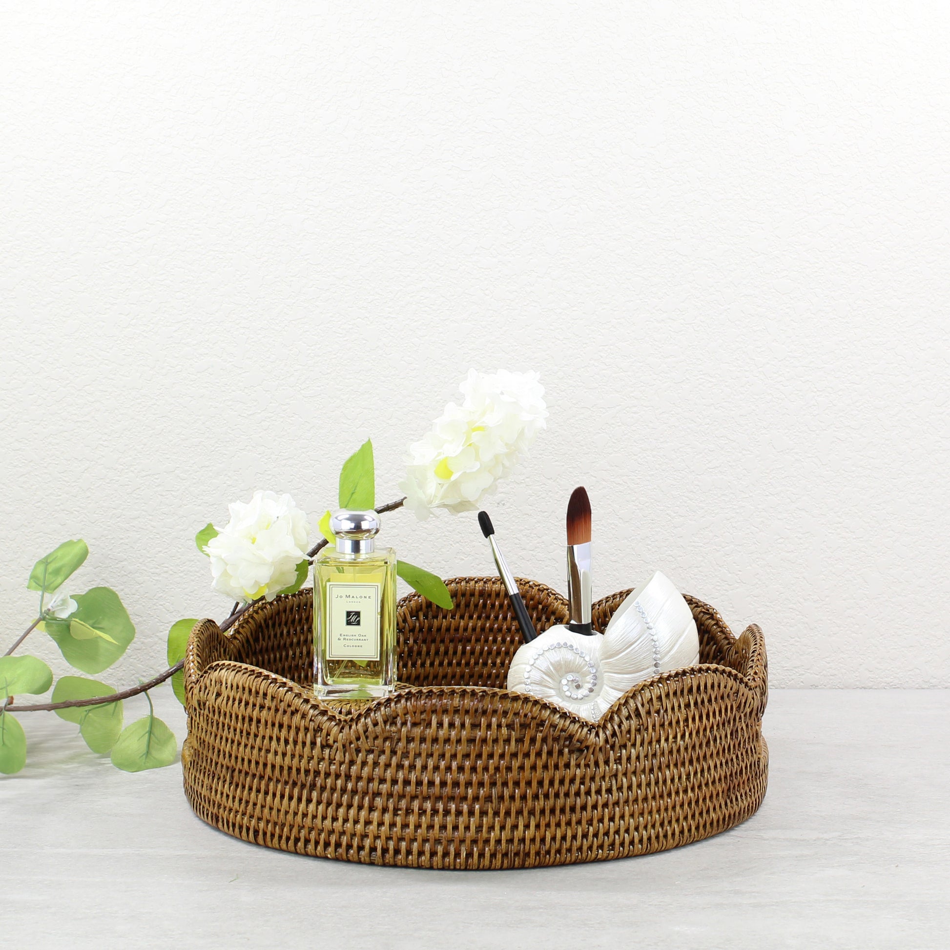 Honey brown scalloped rattan tray with Jo Malone perfume, white flowers, and seashell makeup brush holder for vanity or spa decor.
