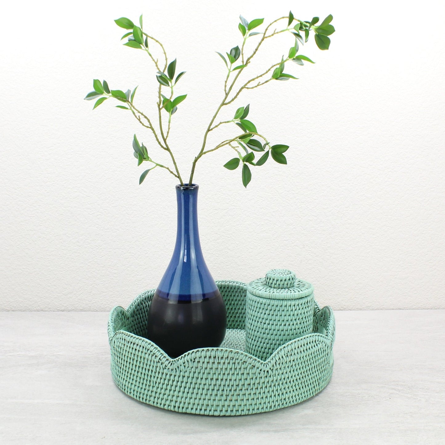 Sage leaf green scalloped rattan tray styled with tall blue vase and matching rattan canister for chic tabletop decor.