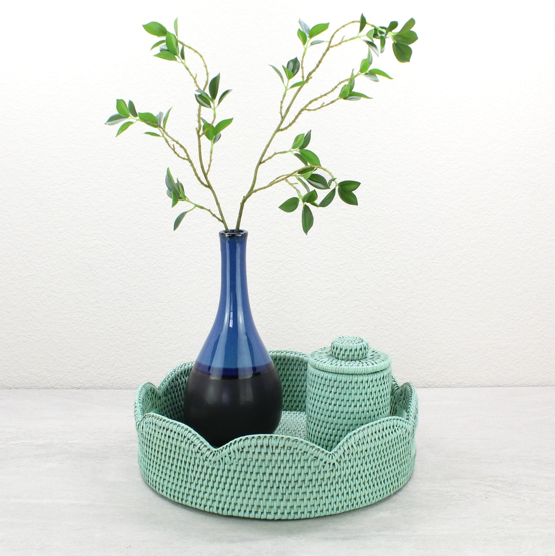 Sage leaf green scalloped rattan tray styled with tall blue vase and matching rattan canister for chic tabletop decor.