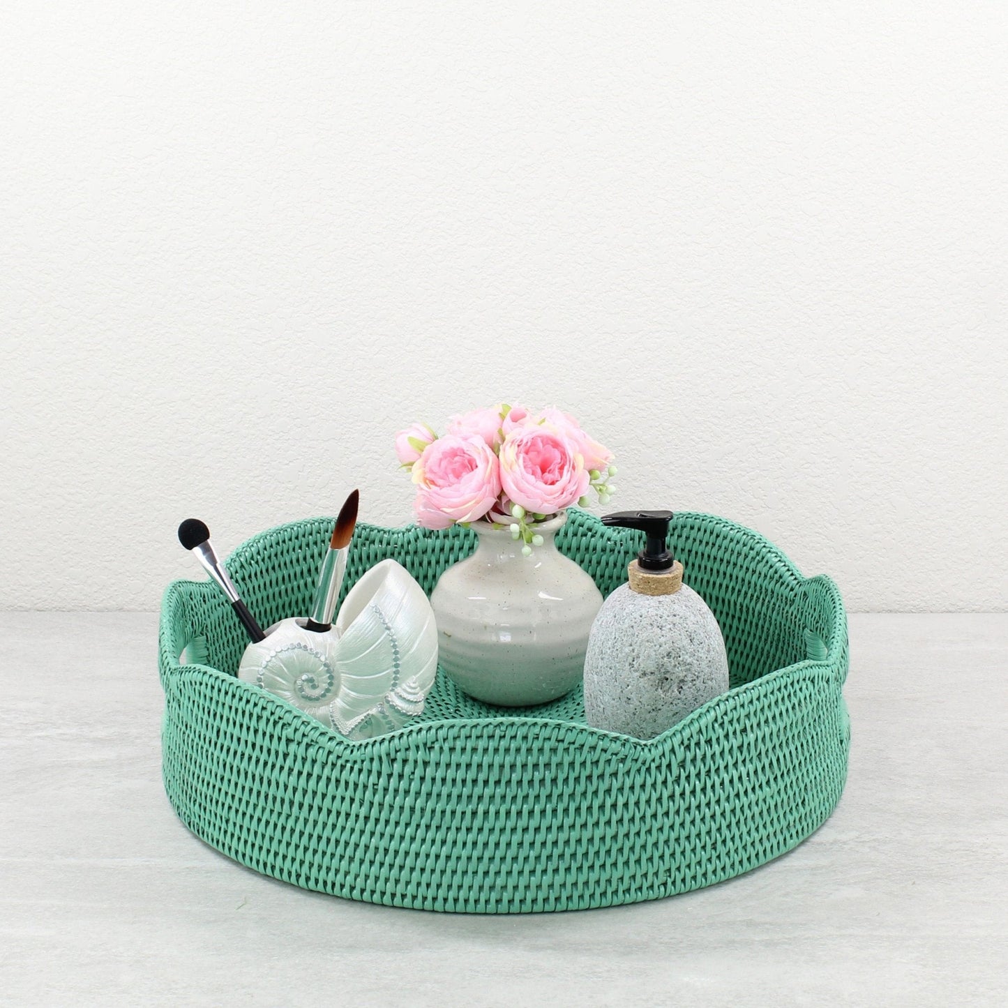 Turquoise scalloped rattan tray styled with pink flowers, soap dispenser, and seashell brush holder for bathroom counter decor.