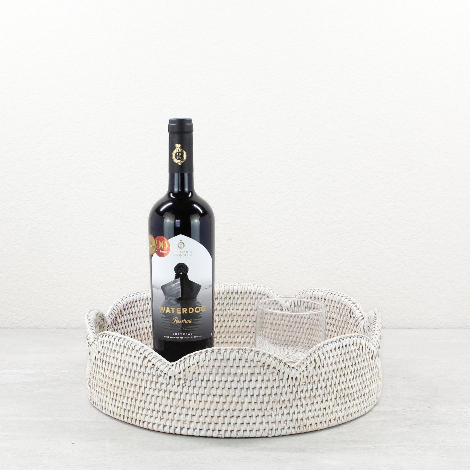 Whitewash scalloped round rattan tray with red wine bottle and glass, styled as a gift set or bar cart accessory.
