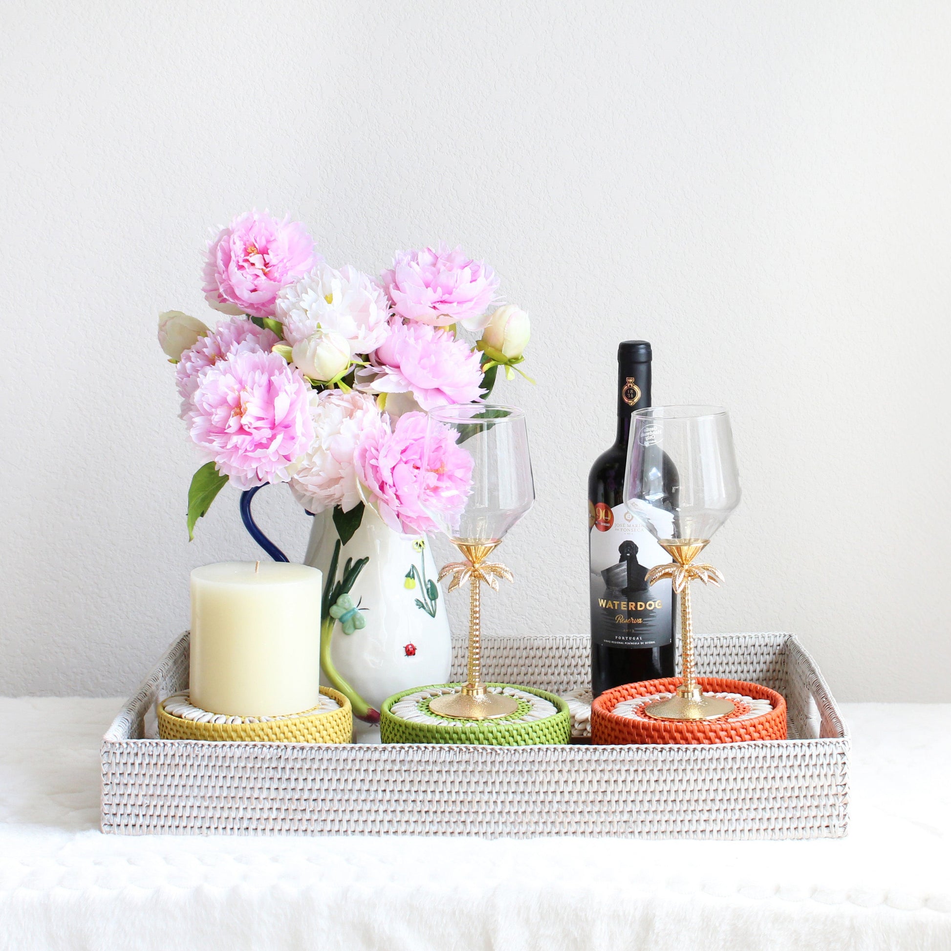 Anniversary gift set featuring a handwoven whitewash rattan tray, seashell coasters, and a hand-painted stoneware pitcher styled with wine glasses and flowers.