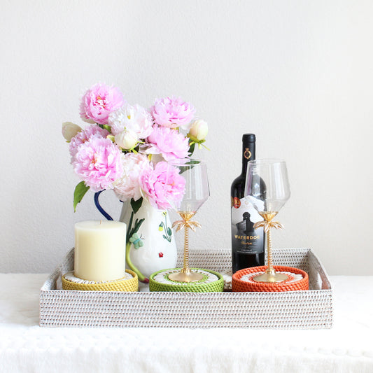 Anniversary gift set featuring a handwoven whitewash rattan tray, seashell coasters, and a hand-painted stoneware pitcher styled with wine glasses and flowers.