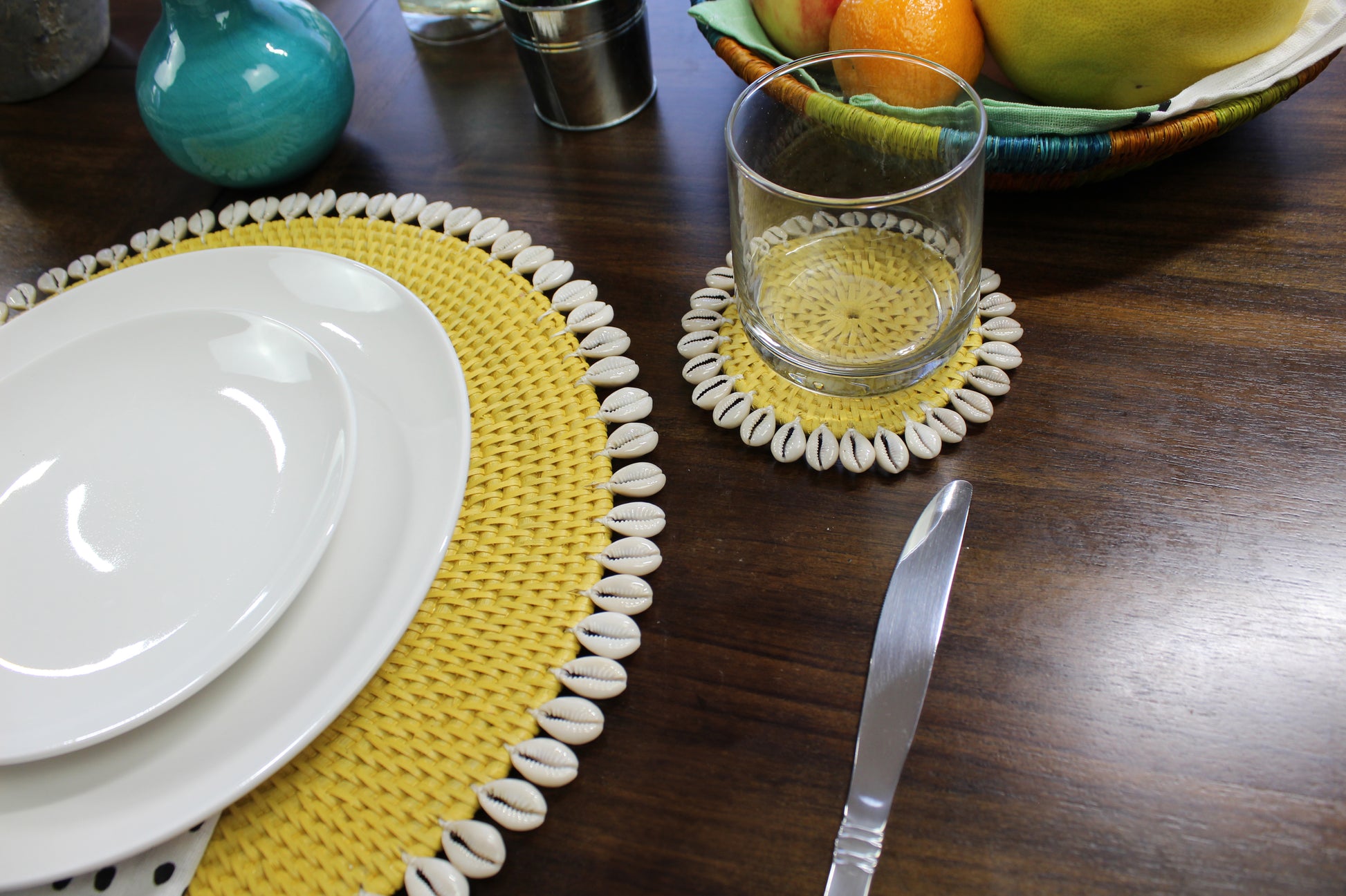 Yellow rattan coaster with seashell trim styled on a wooden dining table with glassware and matching seashell placemat for an elegant coastal-inspired tablescape